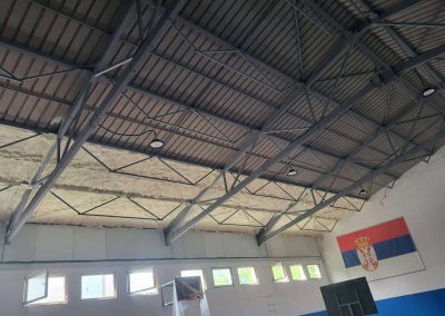 Izolacija sportske hale poliuretanskom penom. Bela izolaciona pena naneta na visoki plafon i gornje delove zidova velike hale, pružajući efikasnu toplotnu zaštitu prostranom sportskom objektu.