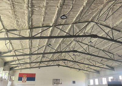 Potpuno izolovani krov sportske hale poliuretanskom penom. Bela izolaciona pena ravnomerno prekriva celokupnu unutrašnju površinu krova, pružajući sveobuhvatnu toplotnu zaštitu i energetsku efikasnost velikom sportskom objektu.
