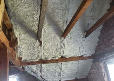 Izolovani krov stambene kuće poliuretanskom penom. Bela izolaciona pena ravnomerno pokriva celokupnu unutrašnju površinu krovne konstrukcije, uključujući grede i prostor između njih. Ova sveobuhvatna izolacija obezbeđuje optimalnu toplotnu zaštitu i povećava energetsku efikasnost domaćinstva.